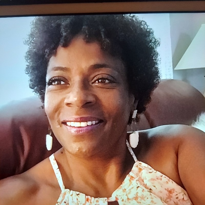Smiling woman with curly hair sitting indoors, wearing a floral top and dangling earrings. The background includes a sofa and a lamp, creating a cozy atmosphere.
